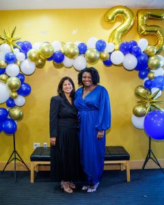 Shanta in a black dress and Aubrey in a blue dress standing under a white, blue, and gold balloon arch.
