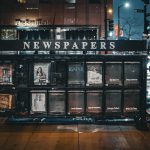 A metal newspaper stand with 2 rows of 13 newspaper containers. Only 5 containers have hardcopy of newspaper.