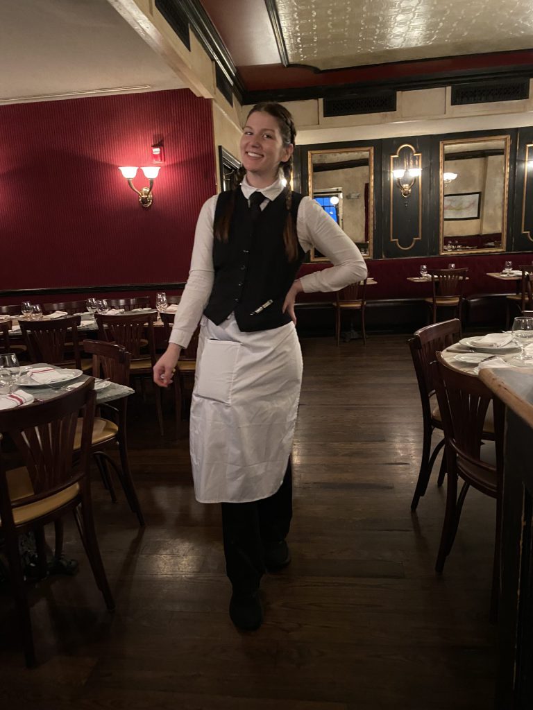 A woman wears a black vest, long sleeve white shirt, white apron, and black pants. Stands in a restaurant dining room