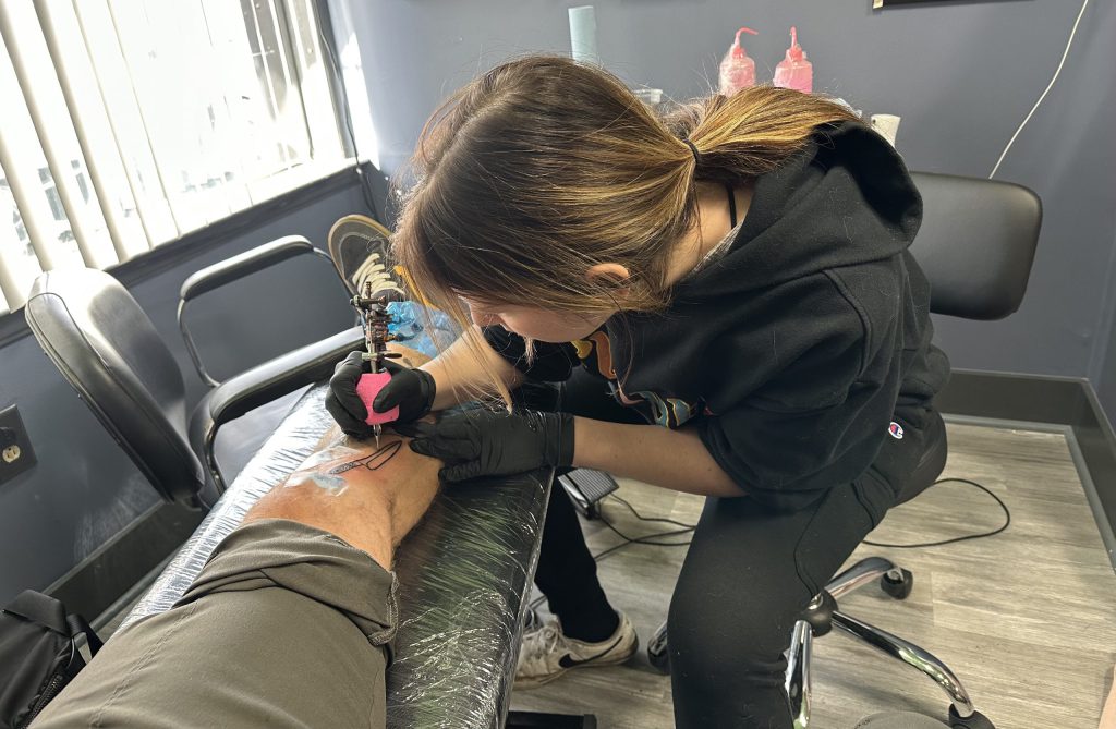Woman tattooing a knife on a mans leg. She wears black pants and hoodie, her hair is in ponytail