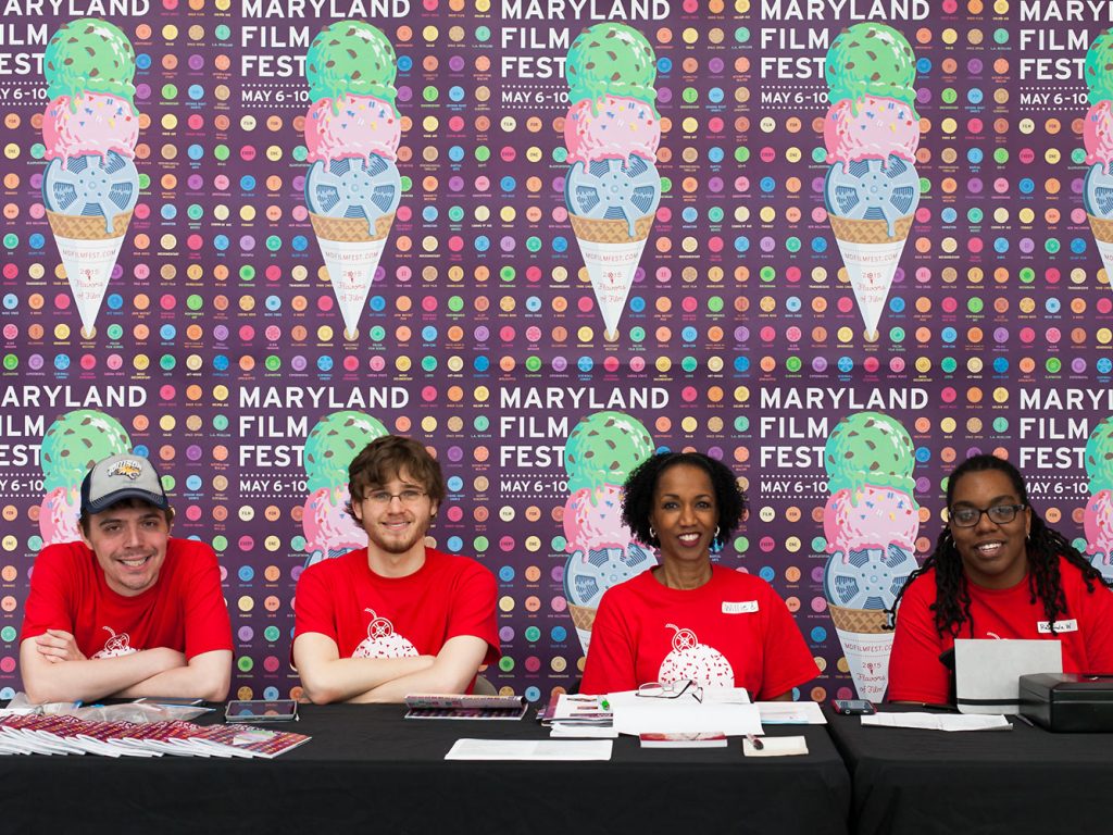 Four images represent different aspects of Post Typography’s branding campaign for the 2015 Maryland Film Festival. Whimsical illustrations of film reels as ice cream cones topped with sprinkles that look like play, pause and fast forward icons reflect the festival’s “2015 Flavors of Film” theme. The poster background features a grid of colorful circle icons representing different genres of film including martial arts, spaghetti western and animation against a dark purple backdrop. The illustrations reference a pop art style by portraying mundane objects, such as film reels, as something else in a colorful way. The overall design aesthetic references modernism because it features a grid and simple sans serif typography. The ice cream reel illustration was used in a poster, billboard and t-shirt design.